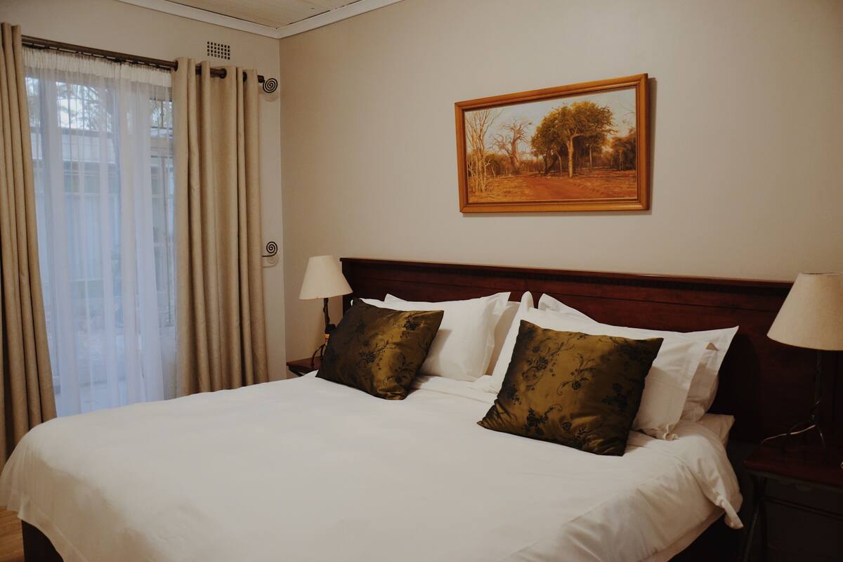A hotel room with a large bed, two bedside lamps, a painting on the wall, and a window with curtains.