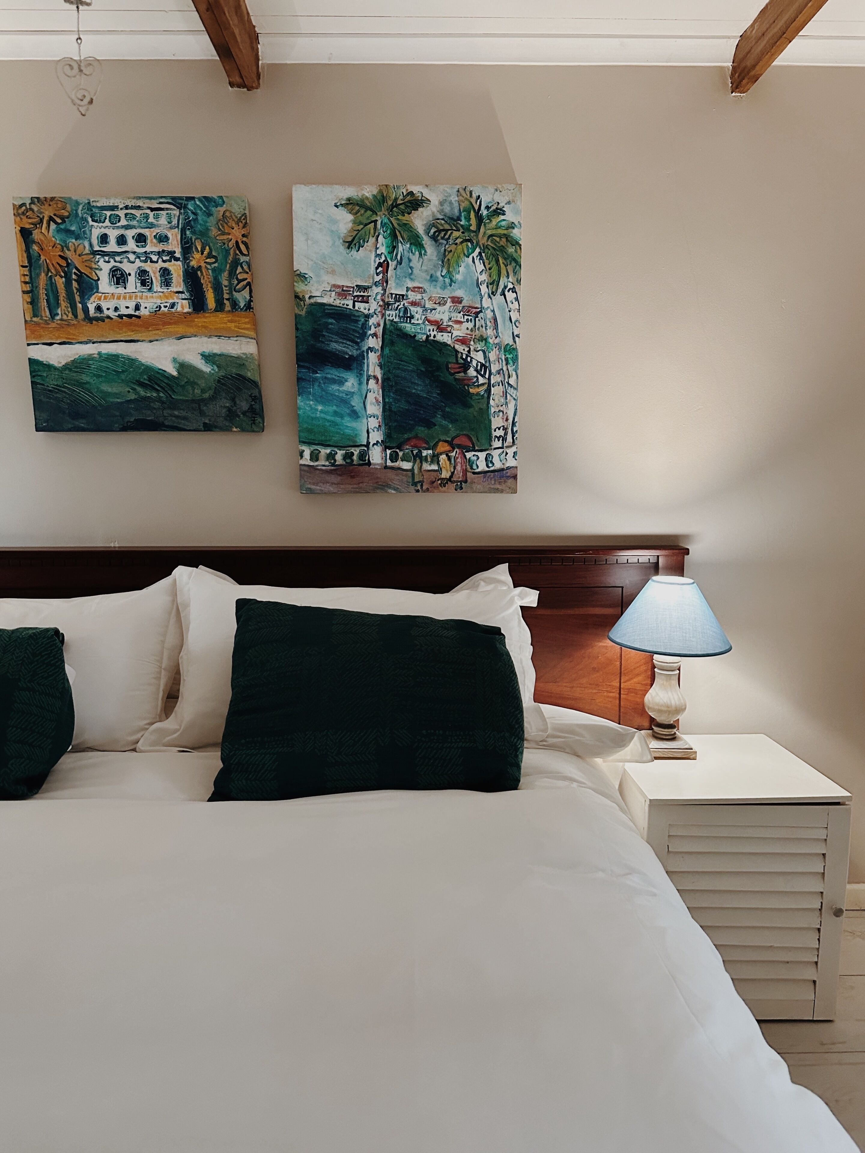 Un dormitorio con cama, almohadas, una mesita de noche, una lámpara y dos cuadros en la pared.