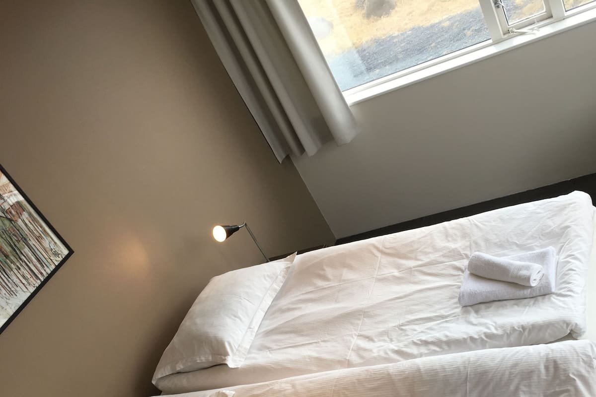 A twin bed with white linens and pillows, a bedside lamp, and a window with curtains.