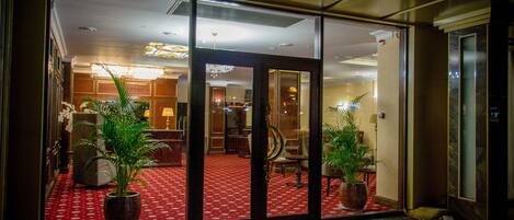 A hotel lobby with a red carpet, glass doors, and potted plants.