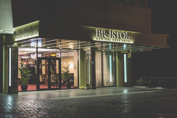 Exterior del Hotel Bristol Central Park de noche, con señalización iluminada, entrada de vidrio y alfombra roja que conduce al lobby.