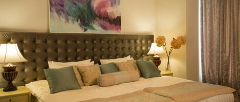 A double bed with a tufted headboard, two bedside tables with lamps, a large abstract painting, and a window with curtains.