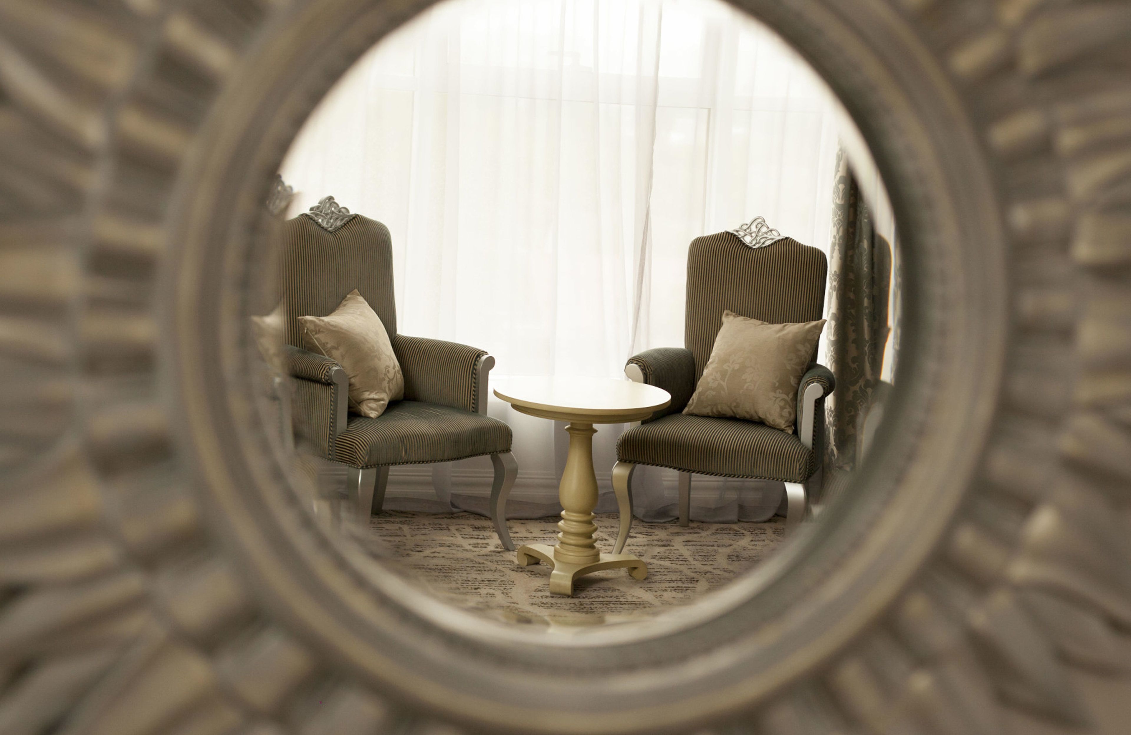 Een ronde spiegel die een kamer weerkaatst met twee fauteuils en een bijzettafeltje.