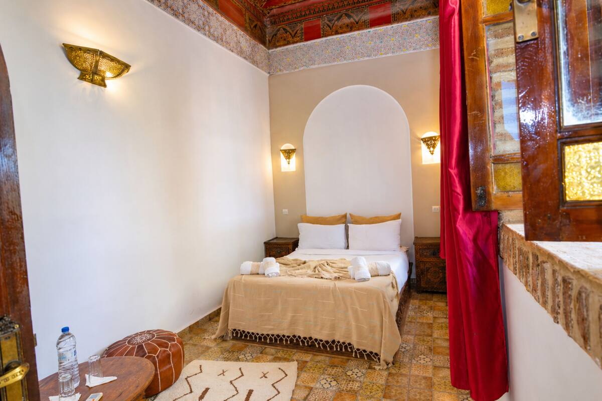 A traditional room with a bed, a small table with a bottle, and a wall-mounted light fixture.