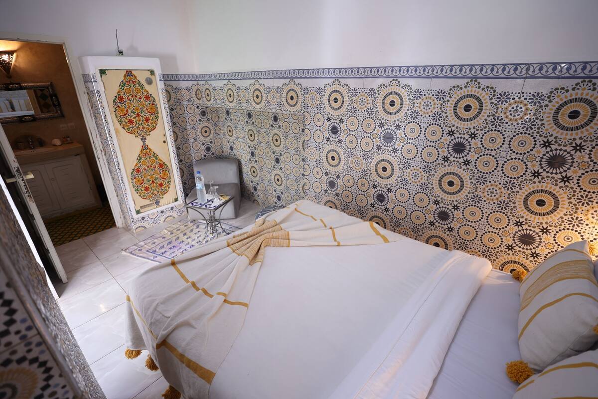 A room with a patterned tile wall, a bed with white bedding and yellow accents, a small table with a lamp, and a door with a mirror.