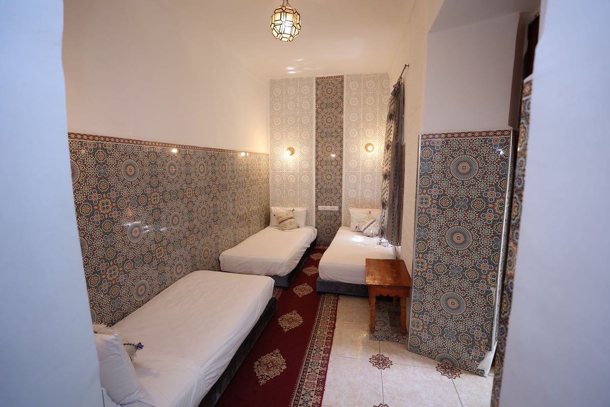 A room with two single beds, a wooden bench, patterned walls, and a hanging light fixture.