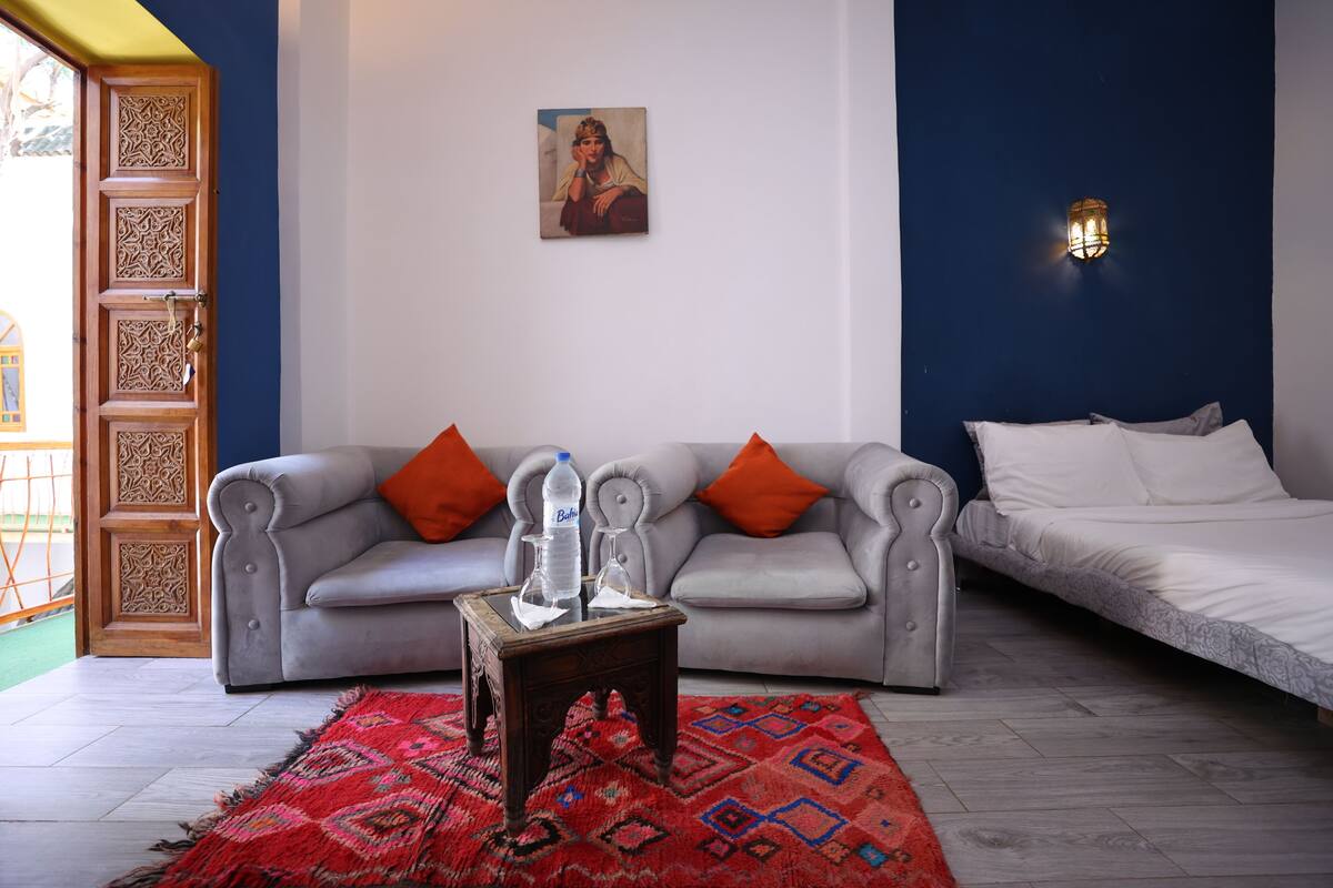 A room with a grey sofa, a bed, a red patterned rug, a small table, and a wall-mounted light fixture.