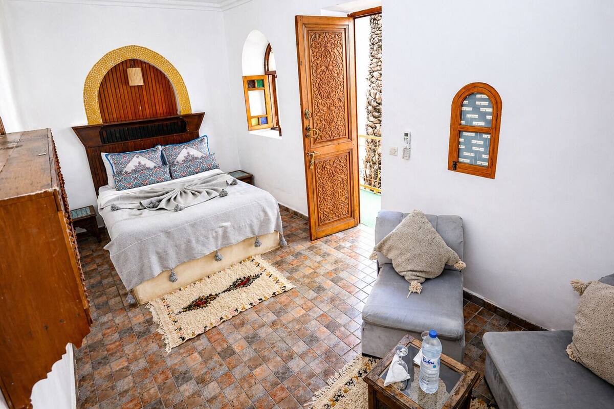 A room with a bed, a sofa, a small table with a bottle, and a door with intricate woodwork.