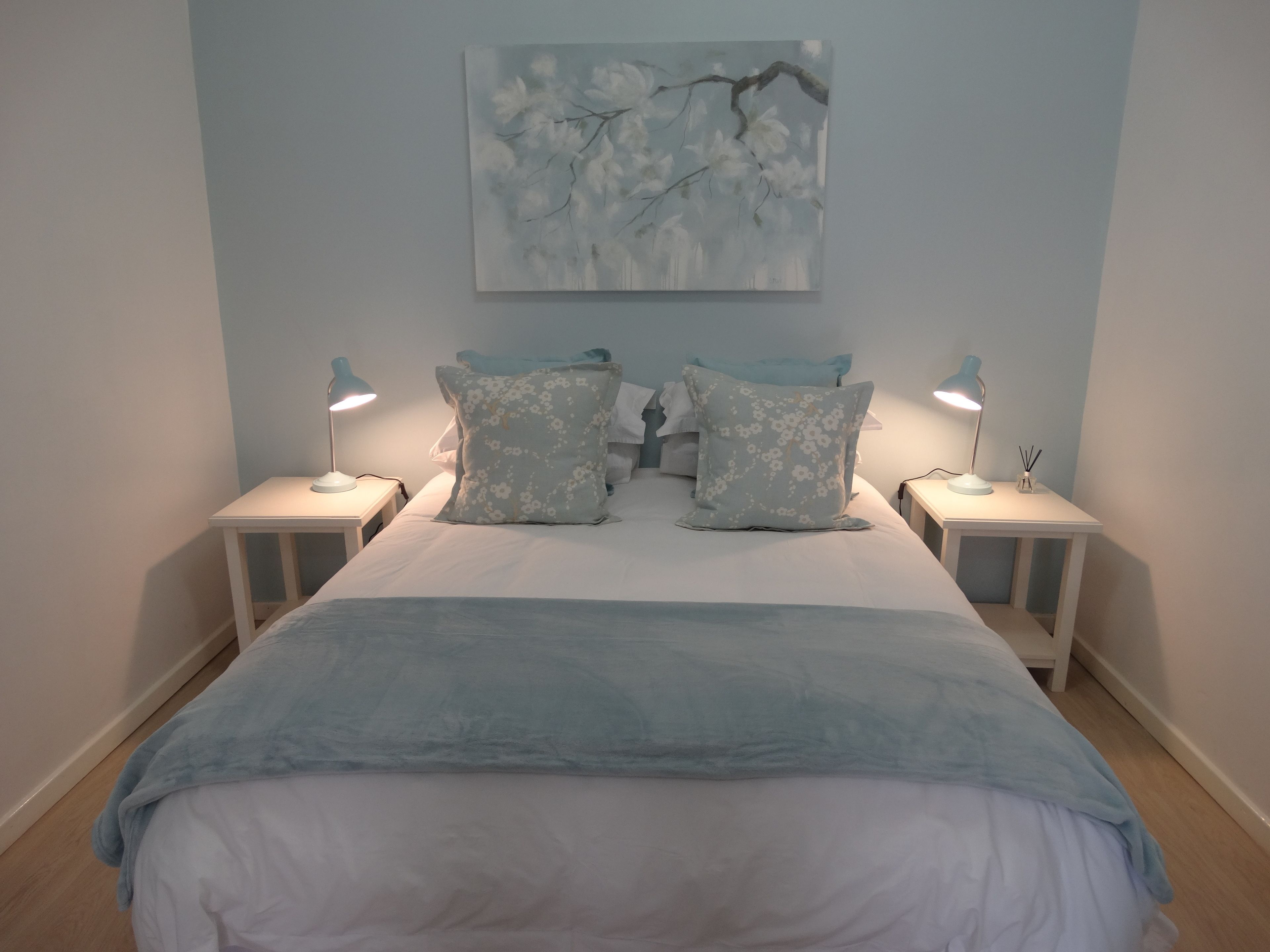 A bedroom with a bed, two bedside tables, two lamps, and a painting on the wall.