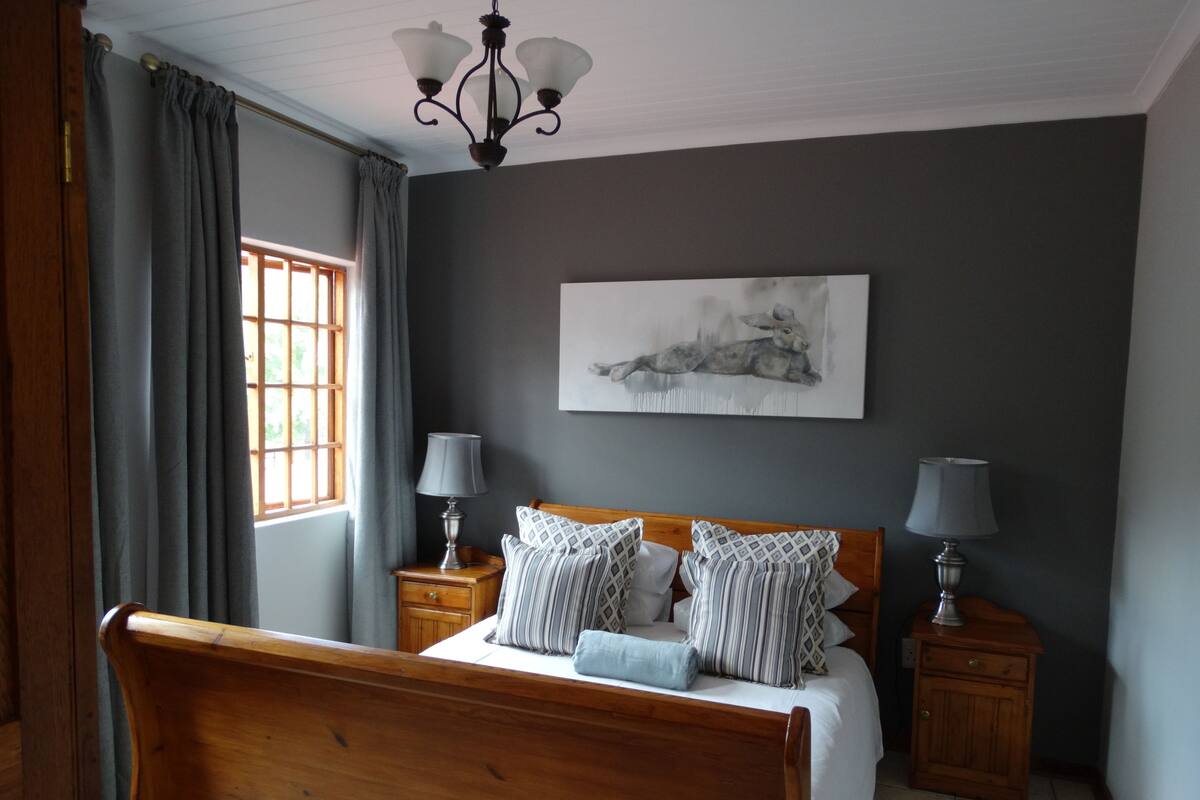 A bedroom with a wooden bed, two bedside tables with lamps, a window with curtains, and a wall-mounted painting.