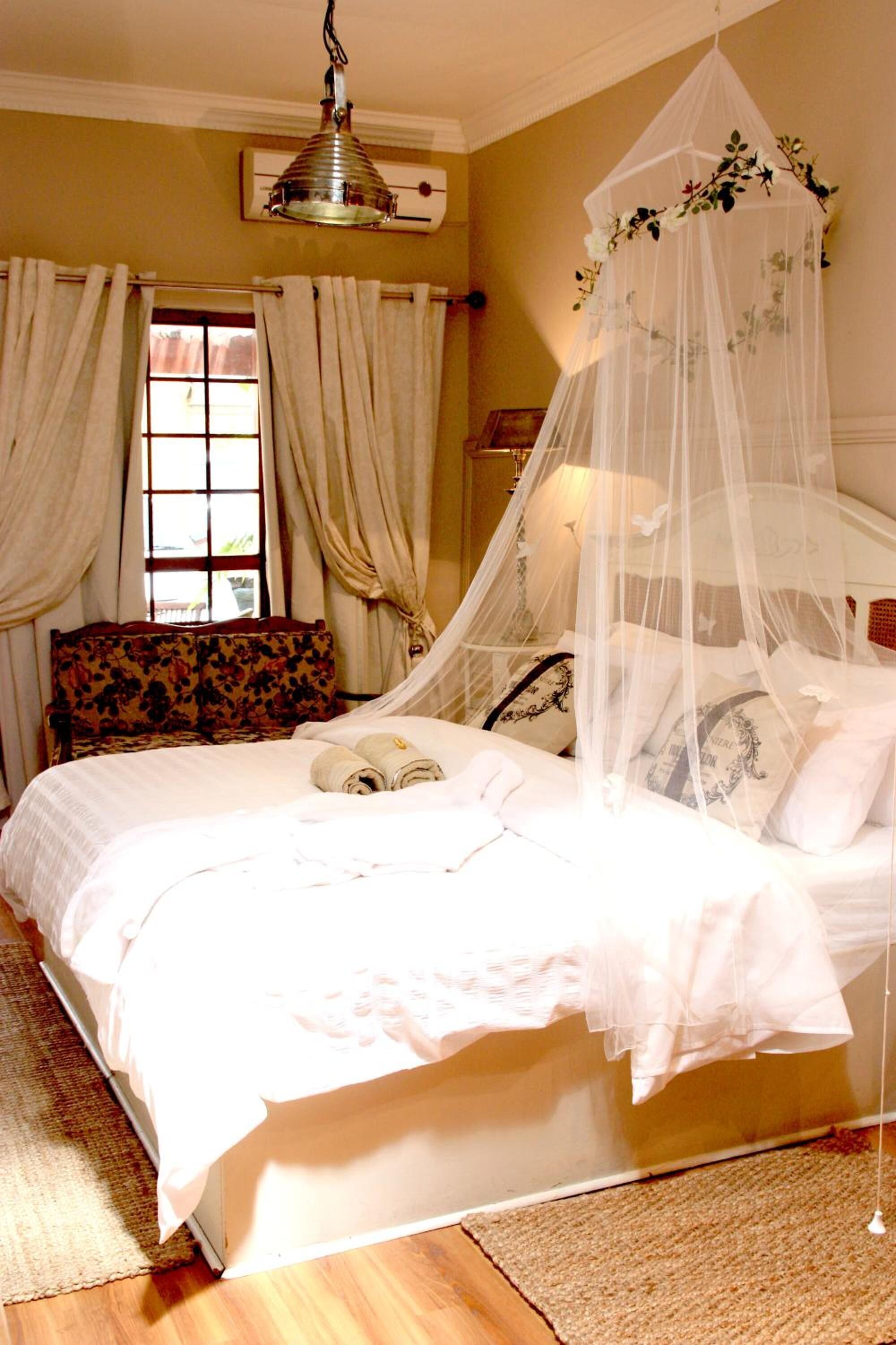 A neatly made bed with a canopy, a floral-patterned armchair, and a window with curtains.