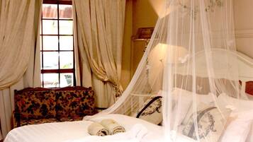A neatly made bed with a canopy, a floral-patterned armchair, and a window with curtains.