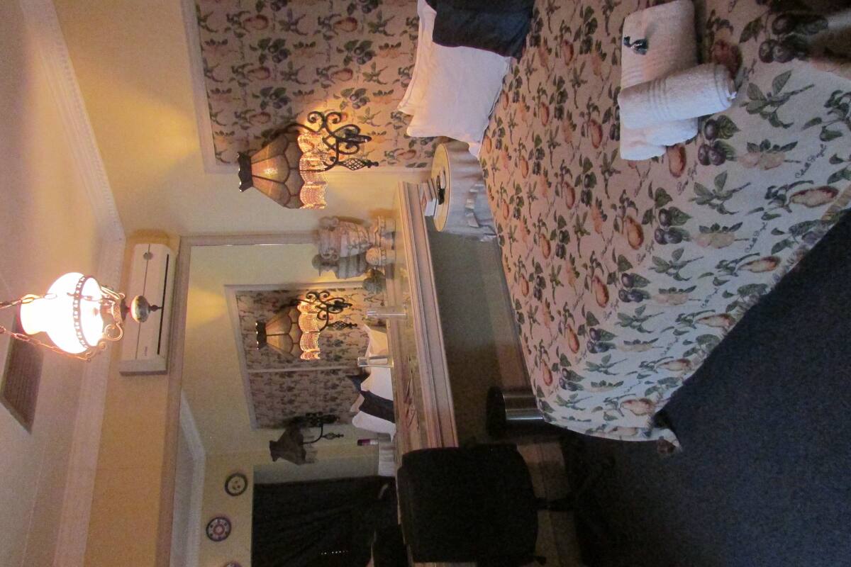 A room with a bed, a desk, and a chair. There is a lamp on the wall and a window with curtains.