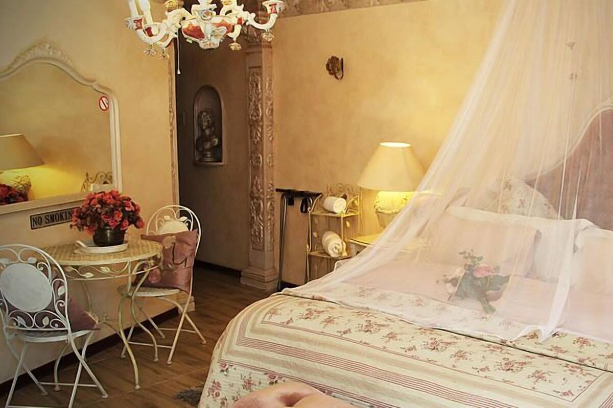 A bedroom with a four-poster bed, a chandelier, a mirror, a desk with a chair, and a nightstand.