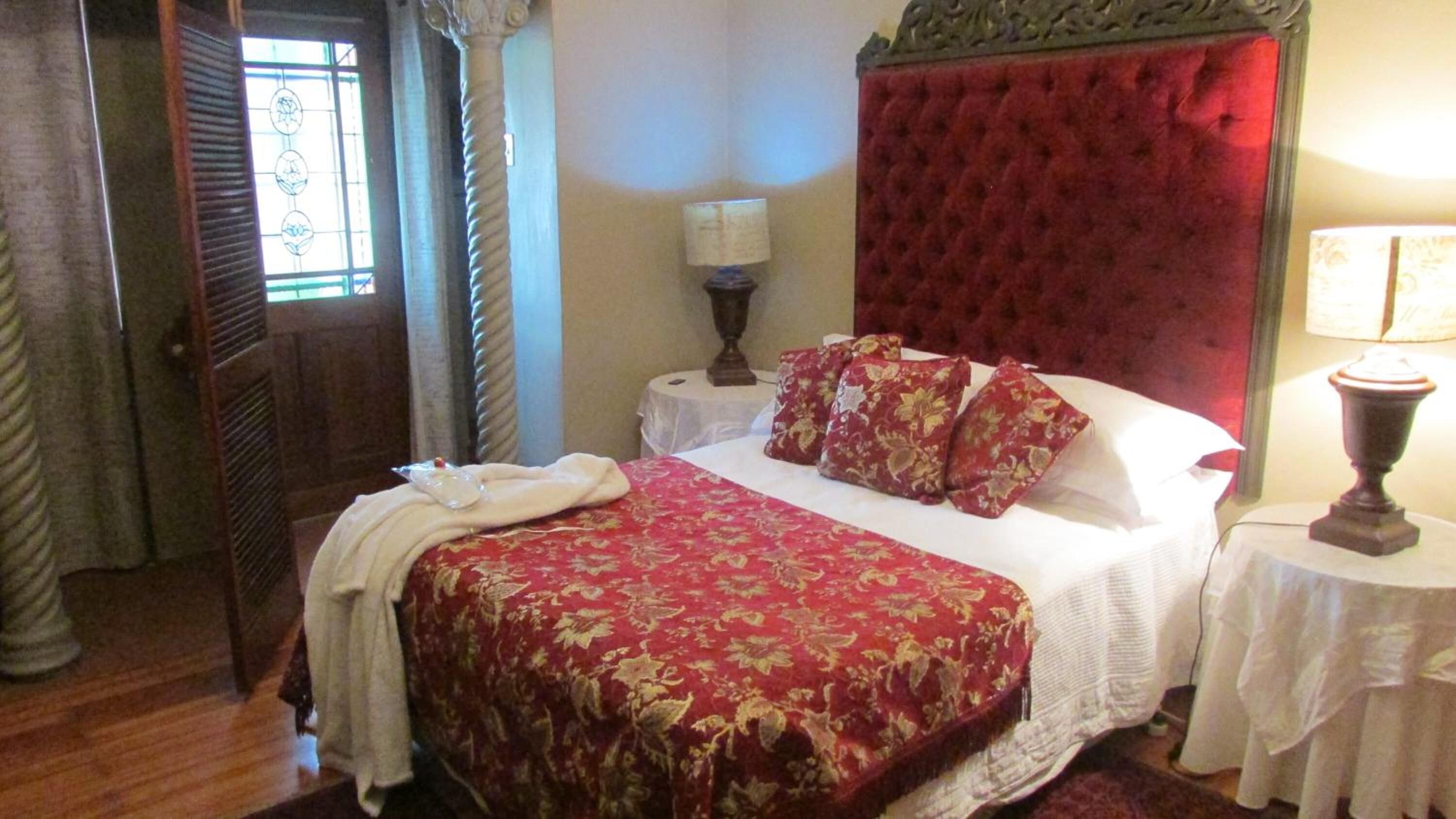 Habitación de hotel con una cama grande, un cabecero tapizado rojo, una lámpara blanca sobre una mesita de noche y una ventana con vistas.