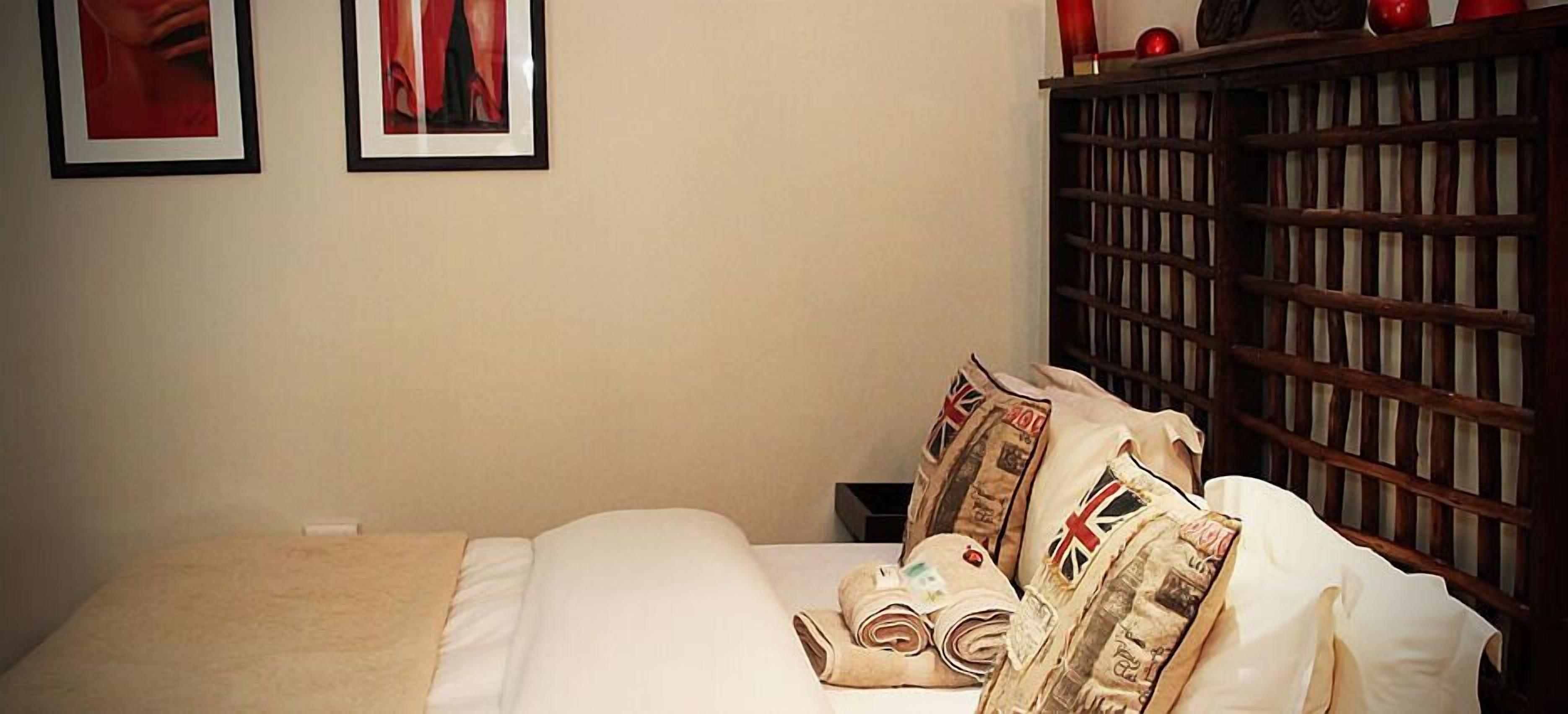 A neatly made bed with white linens and pillows featuring Union Jack designs, a wooden headboard, and a small bedside table with a red object on top.