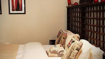 A neatly made bed with white linens and pillows featuring Union Jack designs, a wooden headboard, and a small bedside table with a red object on top.