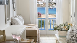 Una habitación acogedora con una cama, una pequeña mesa con un vaso de agua y macarons, una silla de mimbre y vistas al mar y edificios a través de una ventana con marco azul.