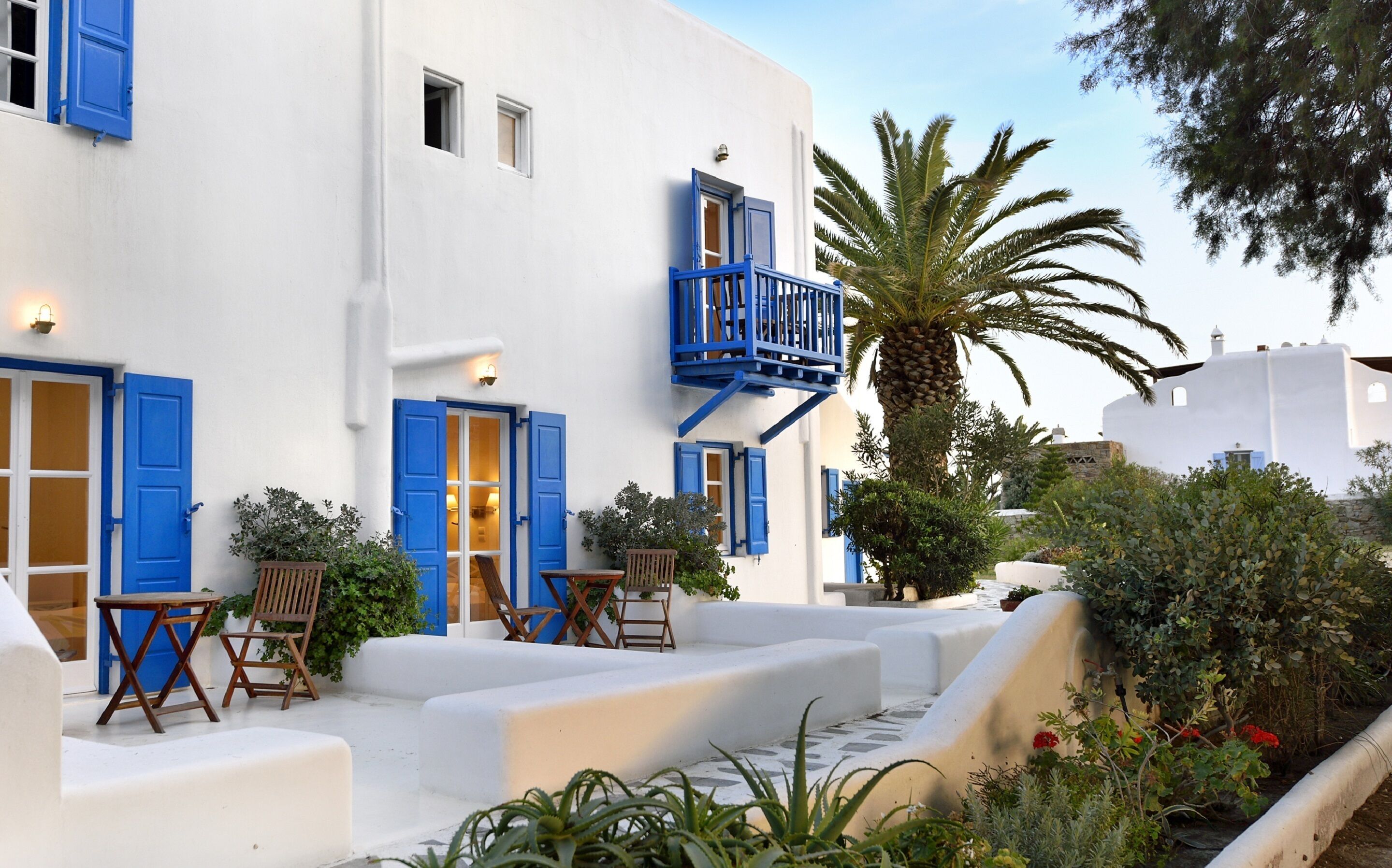 Un edificio blanco con contraventanas azules y un balcón, rodeado de vegetación y palmeras.