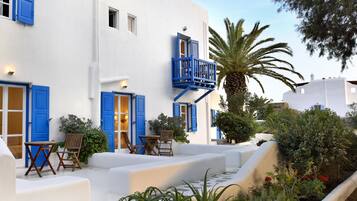 Un edificio blanco con contraventanas azules y un balcón, rodeado de vegetación y palmeras.