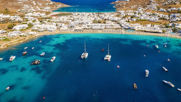 Vista aérea de un pueblo costero con numerosas embarcaciones ancladas en aguas azules cristalinas, rodeado de edificios blancos y un paisaje montañoso al fondo.