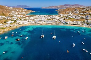 An aerial view of a coastal town with numerous boats anchored in clear blue waters, surrounded by white buildings and a hilly landscape in the background.
