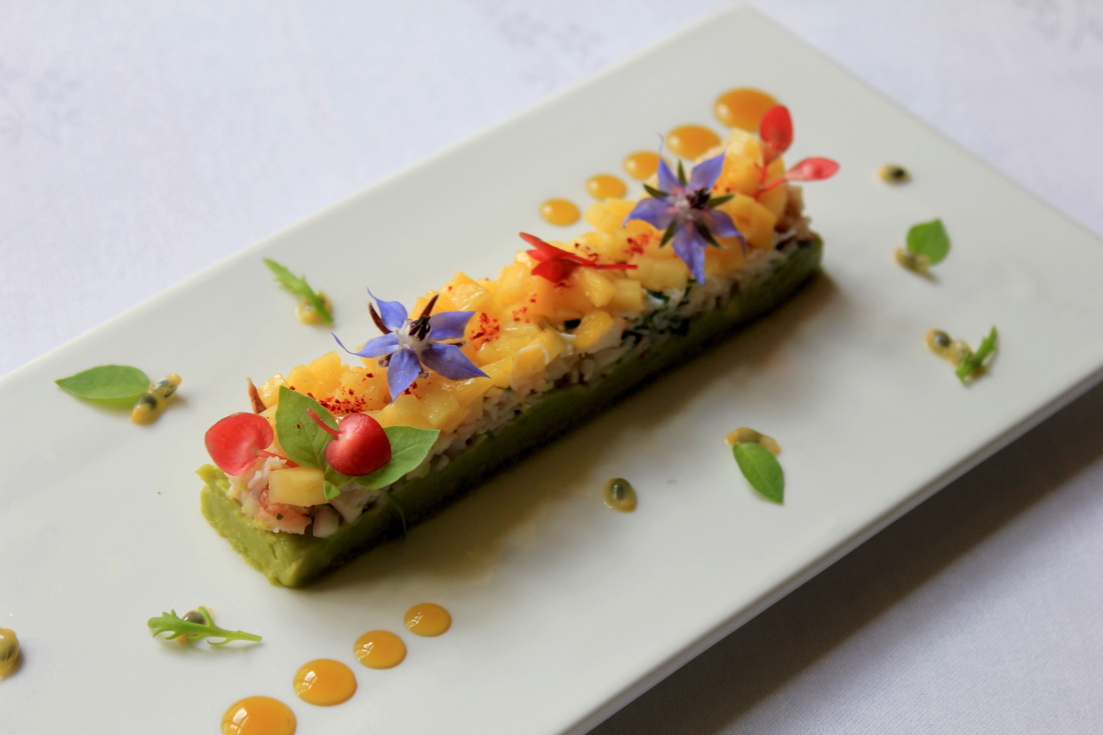 Plato rectangular con un guiso de verdura verde, posiblemente calabacín, cubierto con una mezcla amarilla y roja, adornado con flores y hojas pequeñas.