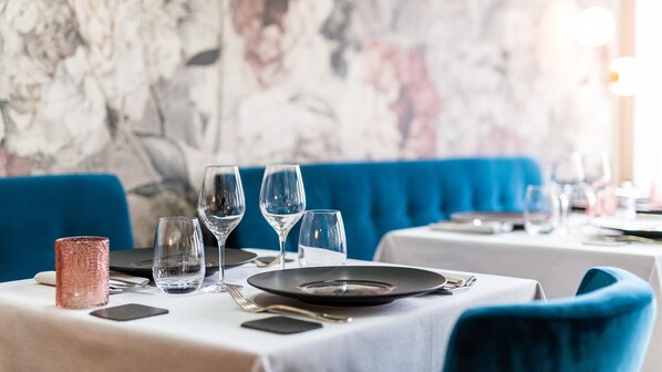 A dining table set with plates, glasses, and cutlery, with a blue upholstered bench and a marble wall in the background.