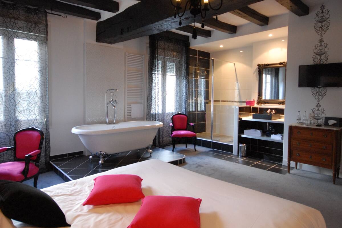 A bedroom with a large bed, a bathtub, a shower, a vanity with a mirror, and a television on the wall.