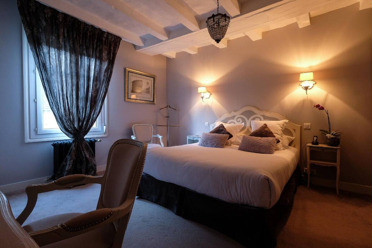 A bedroom with a large bed, two bedside lamps, a chair, a picture on the wall, and a window with curtains.