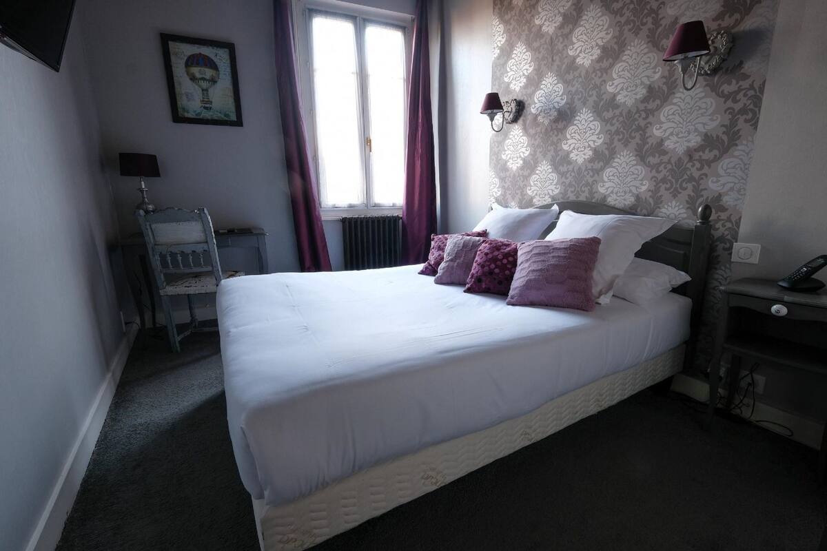 A neatly made bed with white linens and purple pillows, a bedside table with a lamp, and a wall with a patterned wallpaper and a framed picture.