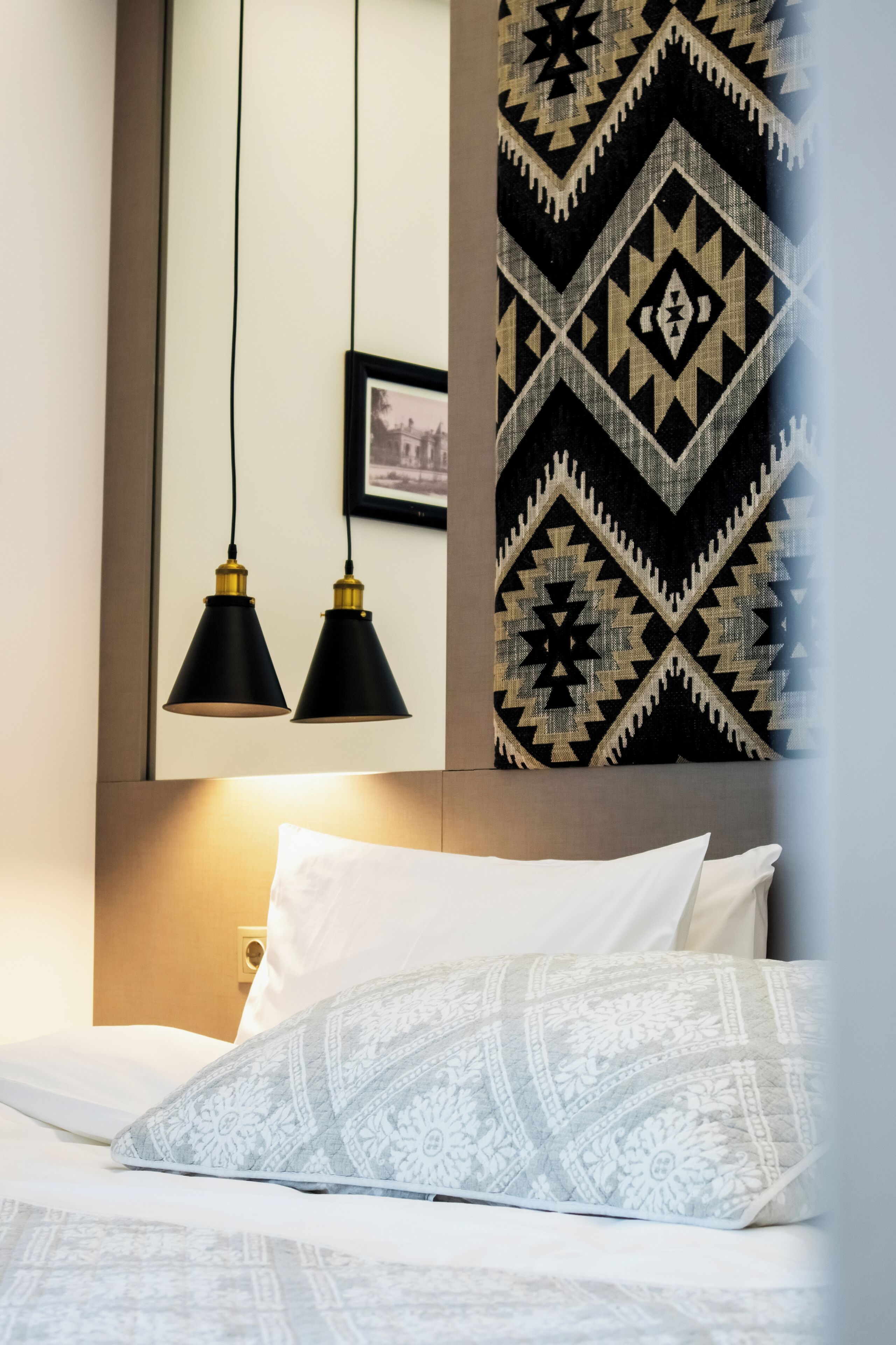 A neatly made bed with white and patterned pillows, a black and white patterned wall hanging, and two black pendant lights.