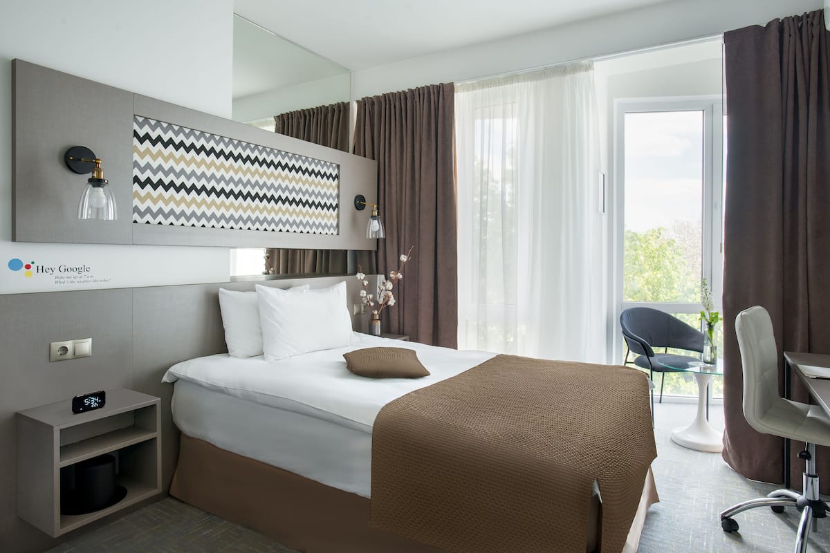 Une chambre d’hôtel moderne comprenant un lit, un bureau, une chaise, une petite table, une horloge murale et un panneau décoratif mural.