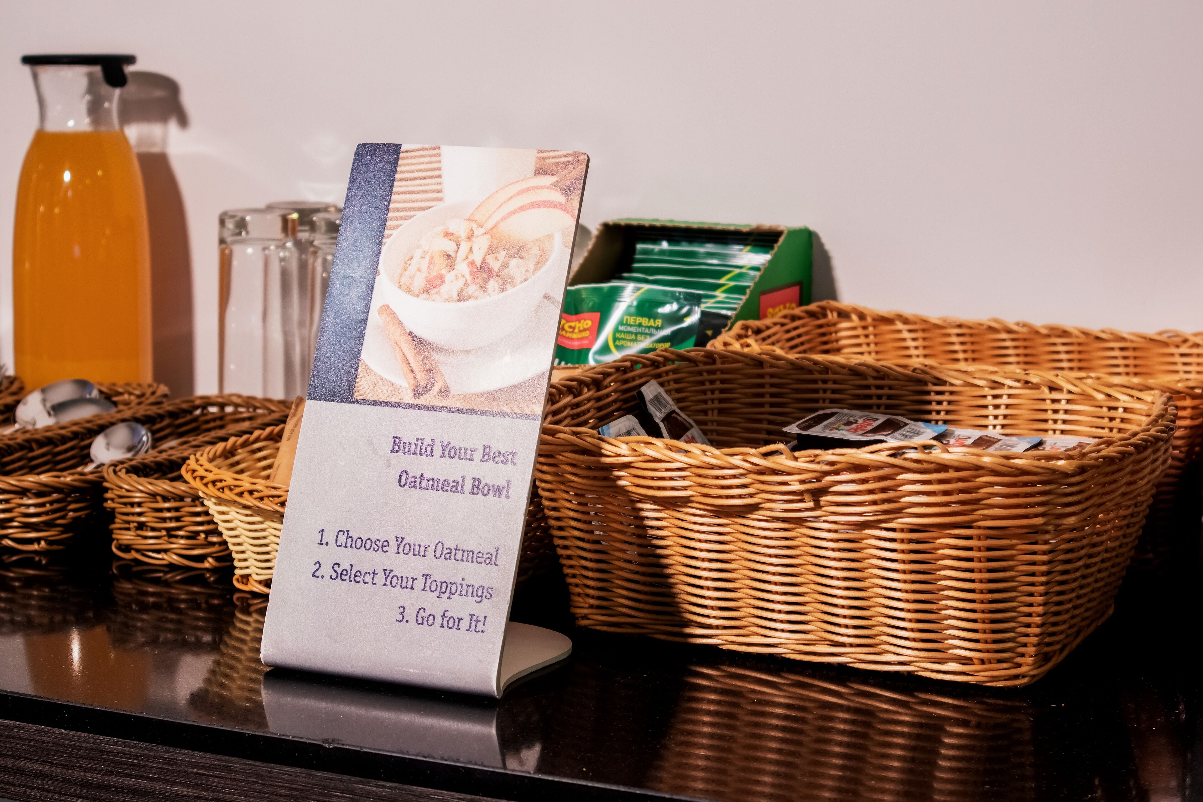 A breakfast setup with a sign instructing to build an oatmeal bowl, a basket of bread, and a bottle of orange juice.