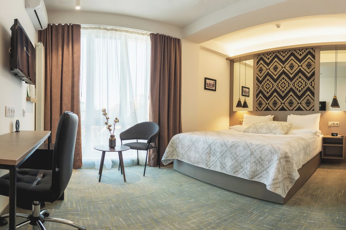 A hotel room with a bed, a desk, a chair, a small table with a vase, and a large mirror on the wall.