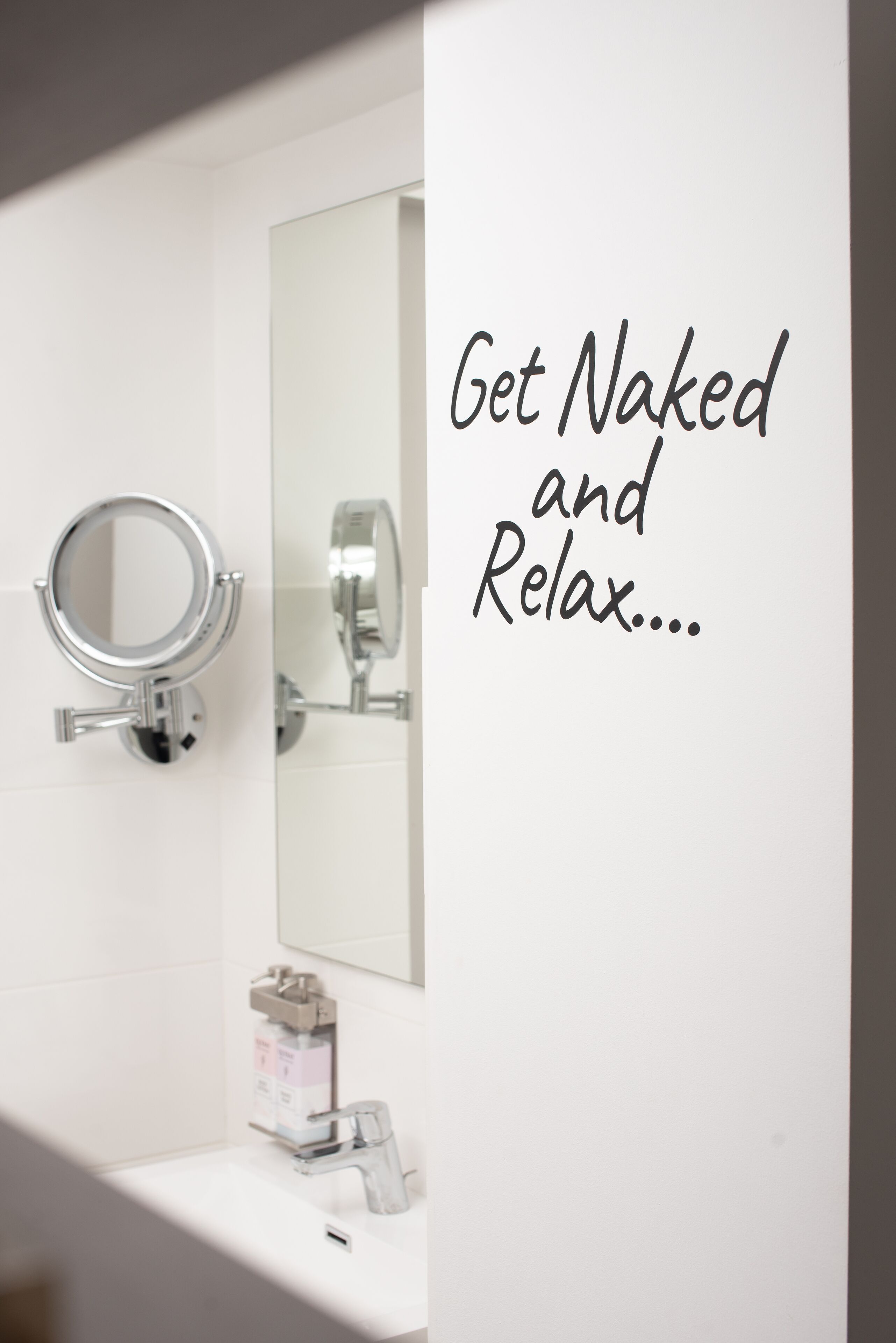 A bathroom with a mirror, sink, and a message on the wall that reads "Get Naked and Relax...".