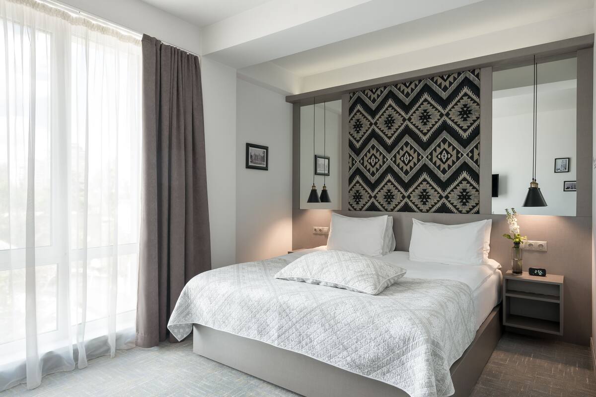 Une chambre d’hôtel moderne avec un grand lit, une table de chevet avec une lampe, un miroir fixé au mur et une fenêtre avec des rideaux.