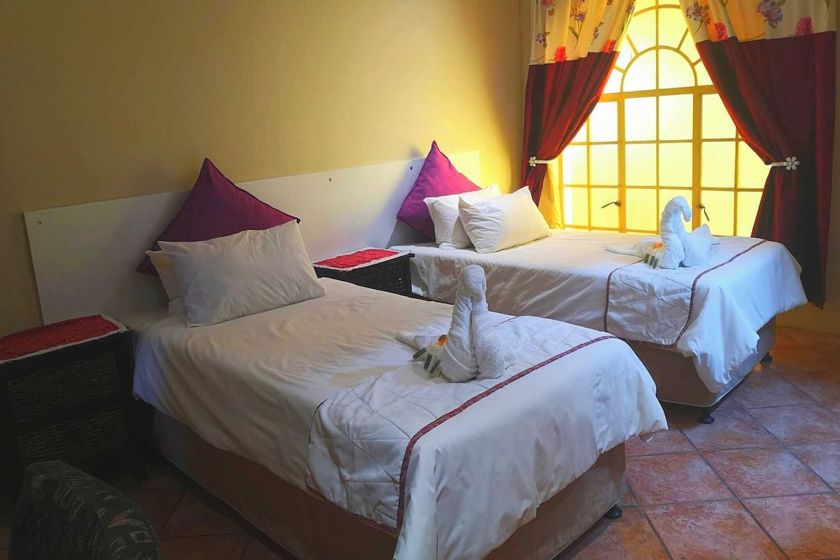 A hotel room with two beds, each with white bedding and red pillows. There is a window with floral curtains and a nightstand with a lamp.