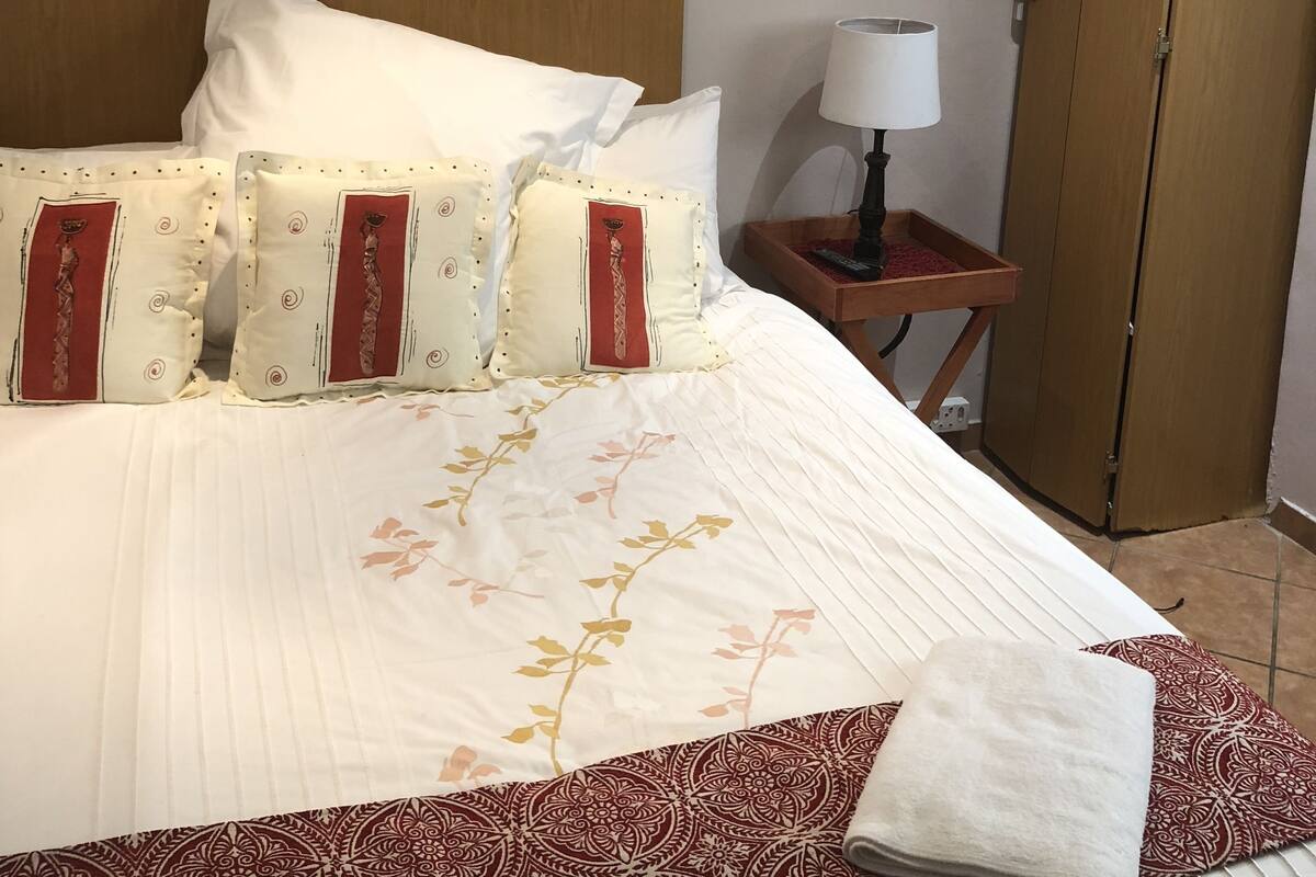 A neatly made bed with a floral-patterned bedspread and decorative pillows, a bedside table with a lamp, and a wooden wardrobe.
