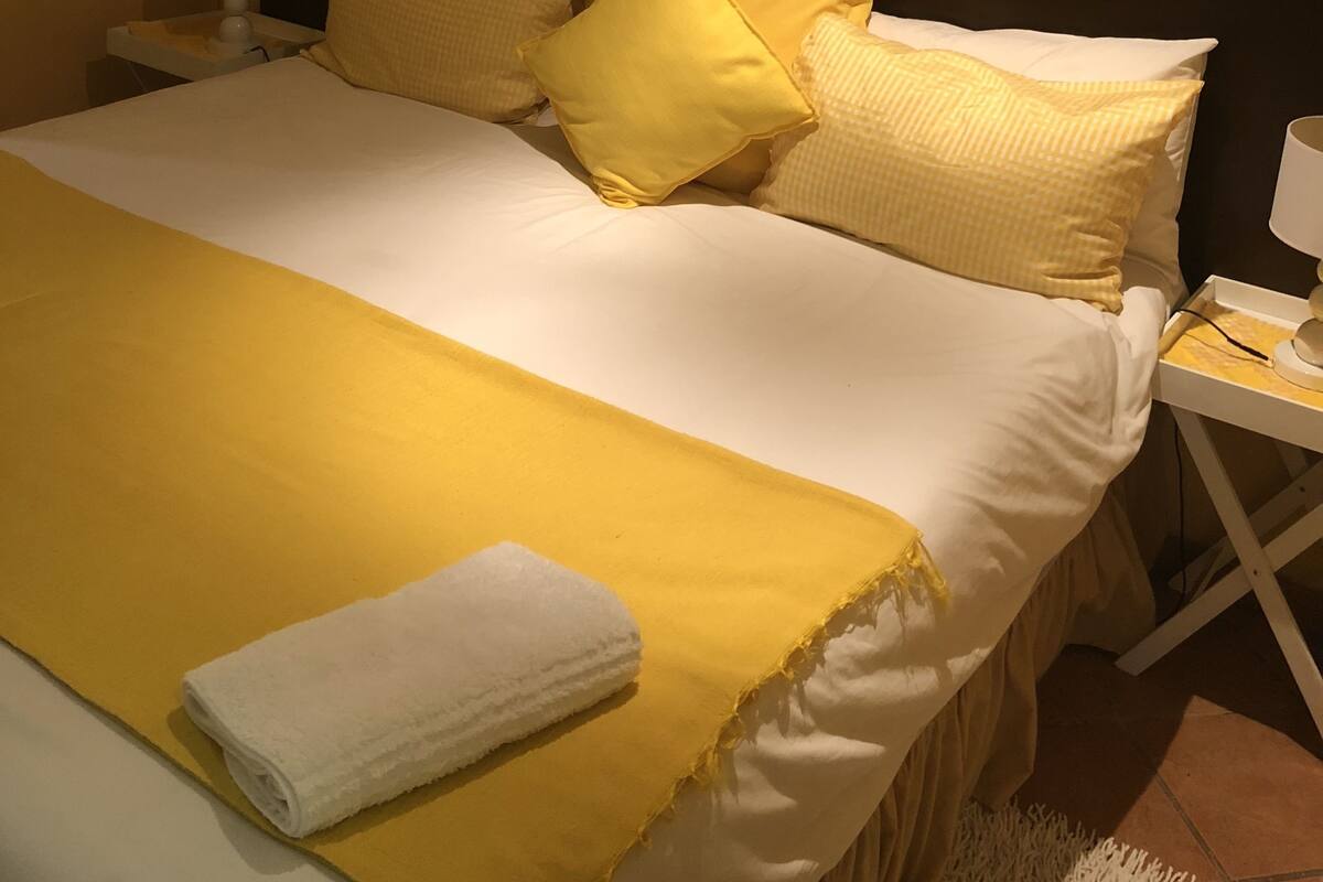 A neatly made bed with a yellow bedspread and pillows, a white towel, and a nightstand with a lamp.