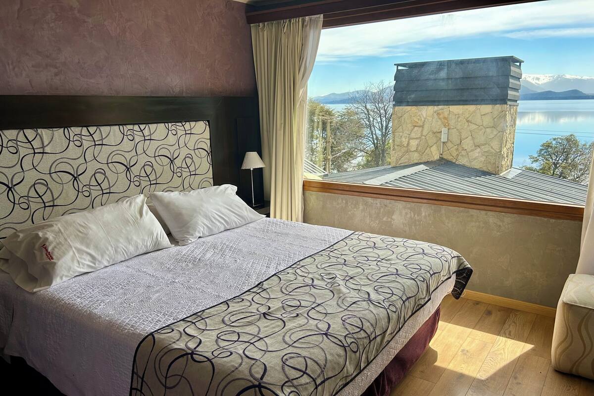 Habitación de hotel con una cama grande, vista a las montañas y al agua, y un balcón con pared de piedra.