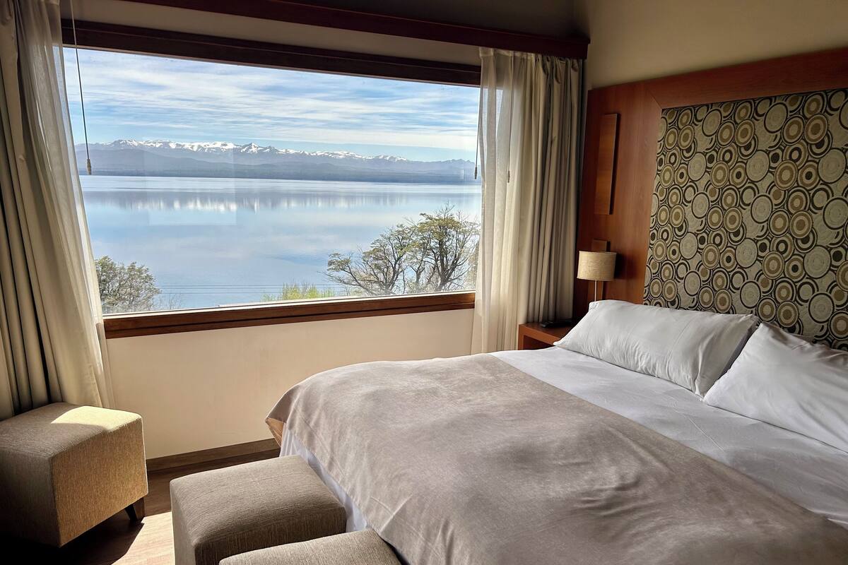 Una habitación de hotel con una cama grande, una mesita de noche, una lámpara y vistas a un lago con montañas al fondo.