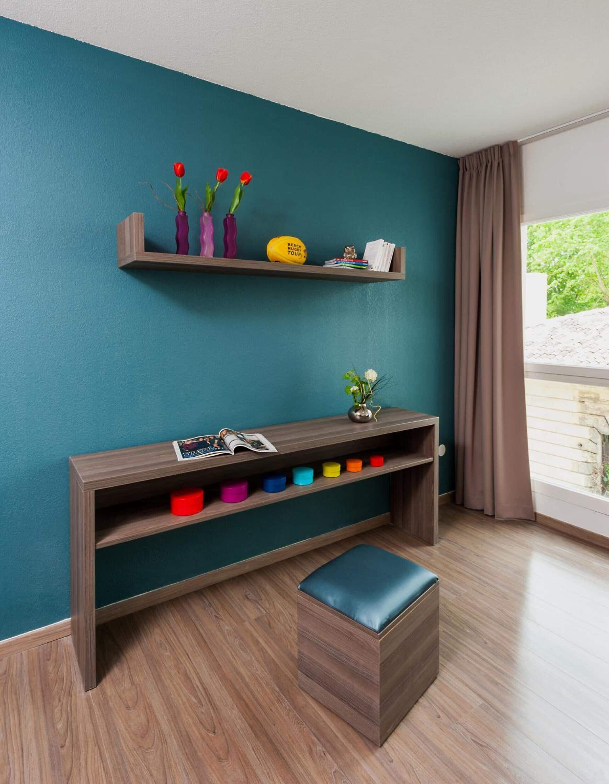Un salon moderne aux murs de couleur turquoise, avec un banc en bois doté de boîtes de rangement colorées, et une petite table sur laquelle sont posés un vase et un livre.