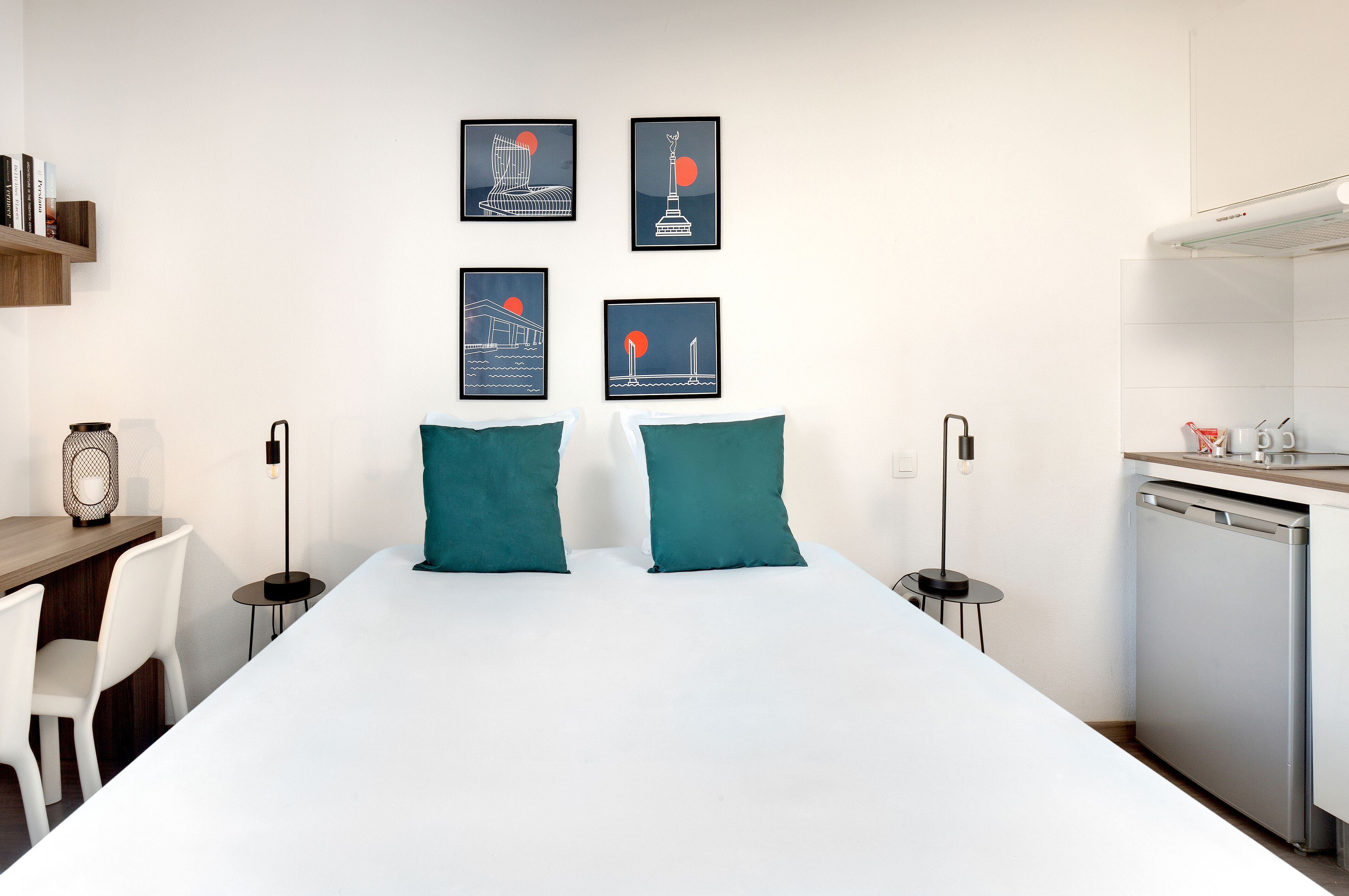 A modern hotel room with a white bed, teal pillows, a desk with a lamp, and a kitchenette.
