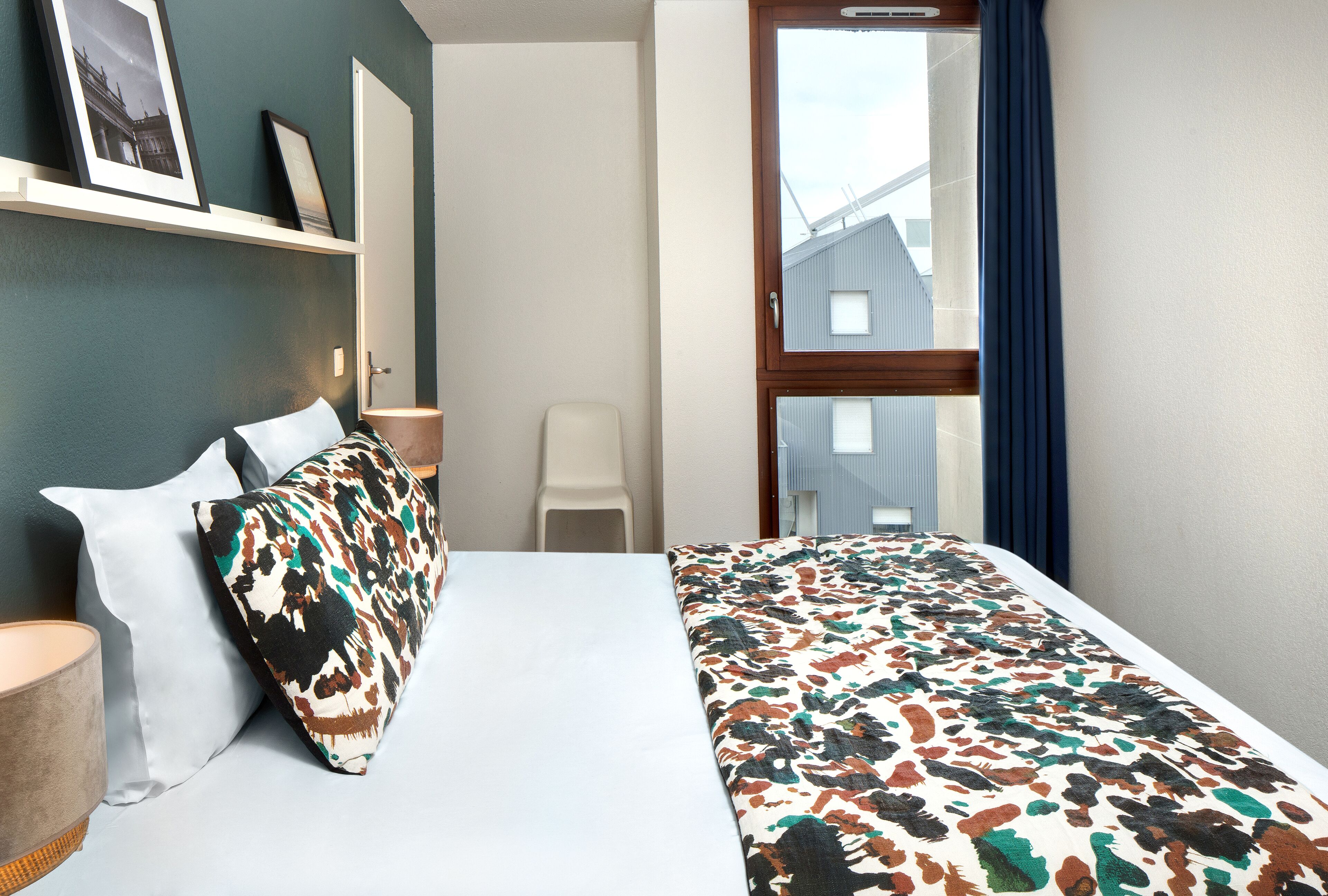 Habitación de hotel con cama, almohadas, mesita de noche, lámpara, silla y ventana con cortinas.