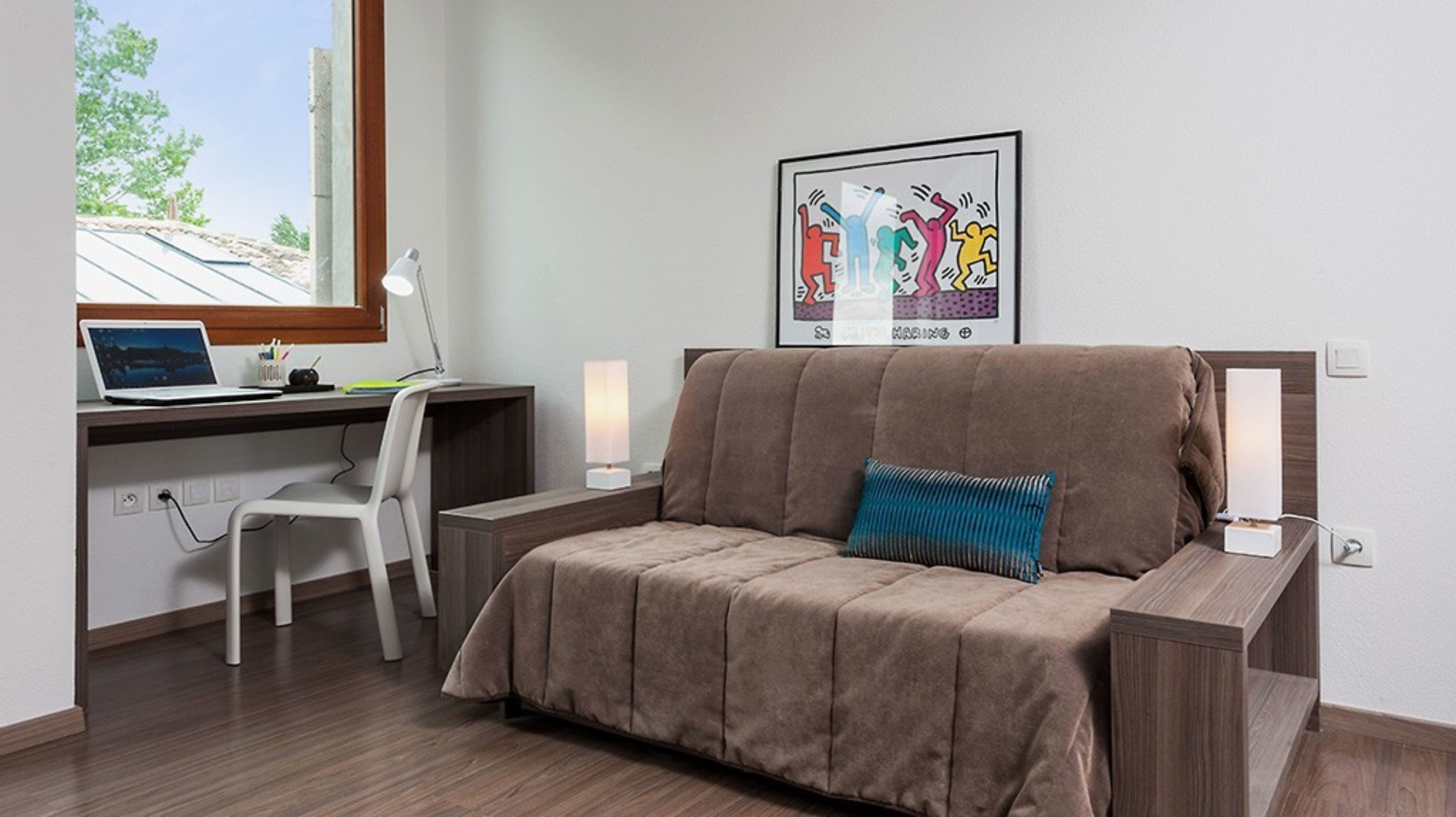 A modern room with a brown sofa, a desk with a laptop, a chair, and a framed artwork on the wall.