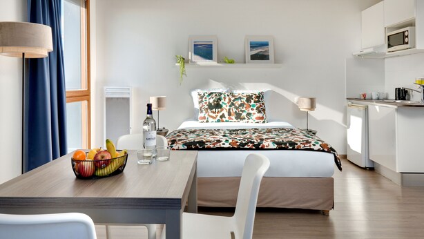 Habitación de hotel moderna con cama, mesa de comedor con fruta, zona de cocina y obras de arte en la pared.