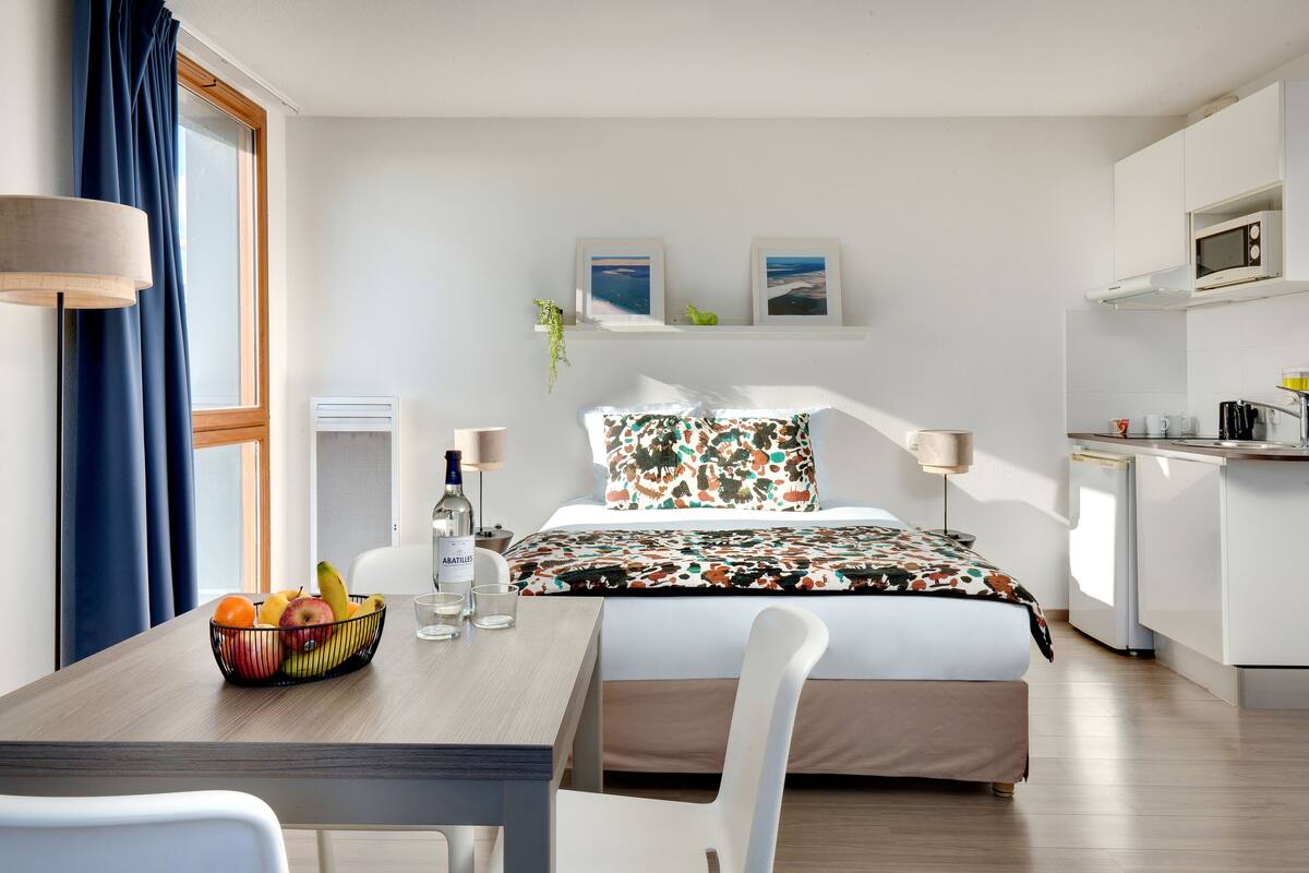 A modern hotel room with a bed, a dining table with fruit, a kitchen area, and artwork on the wall.