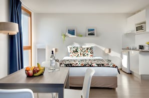 Habitación de hotel moderna con cama, mesa de comedor con fruta, zona de cocina y obras de arte en la pared.