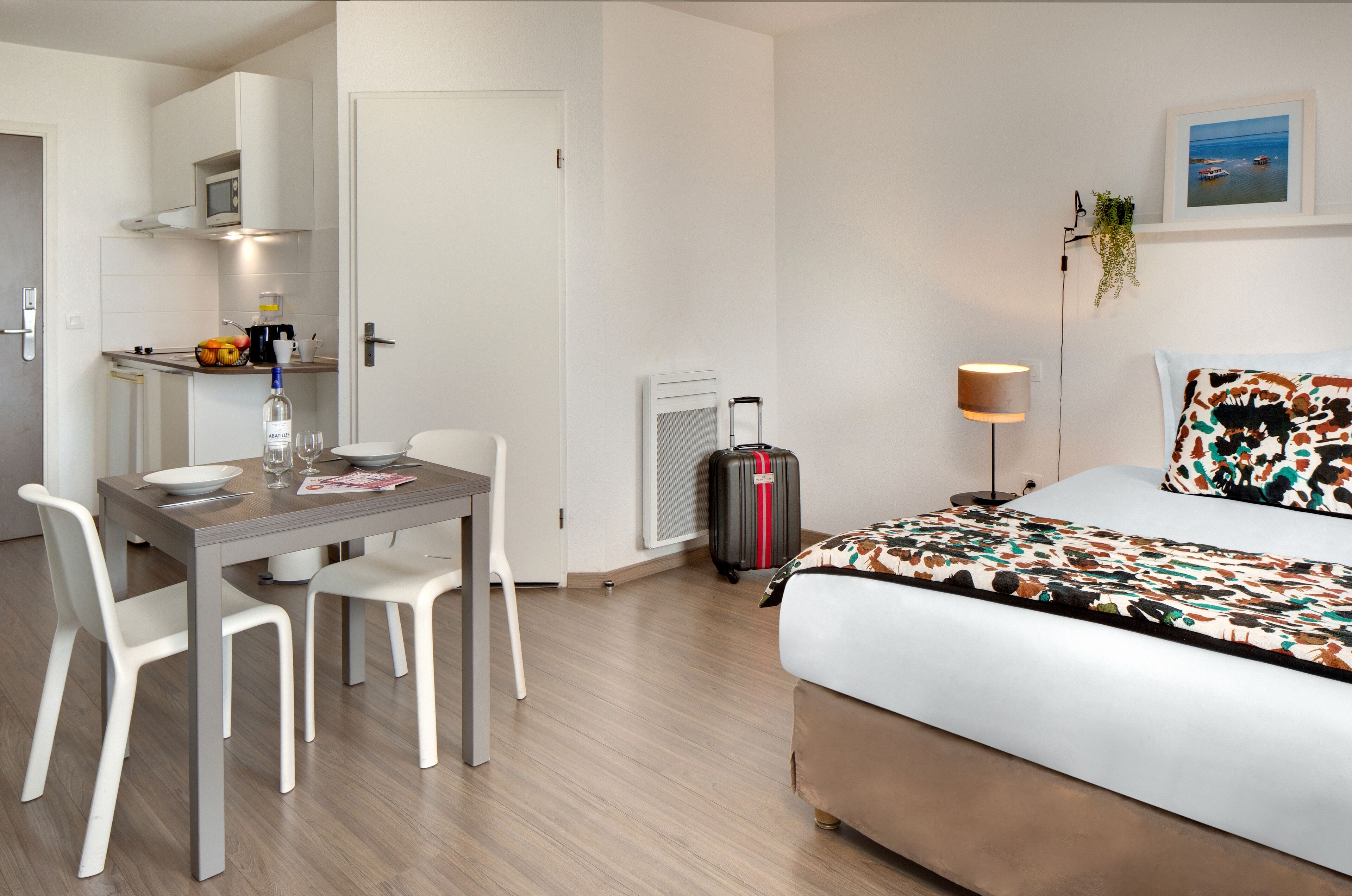 Ein Hotelzimmer mit einem Bett, einem Schreibtisch mit Stühlen, einem Koffer, einer Lampe und einer kleinen Küchenzeile.